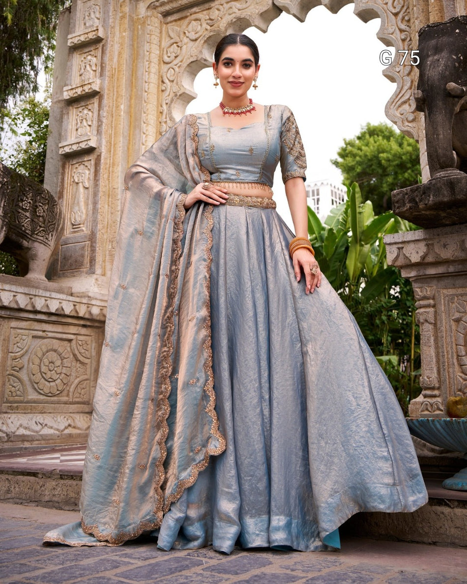 A woman models a traditional light blue anarkali gown with a matching dupatta, featuring gold embroidery, a gold belt, and a square neckline, posed in front of an ornate stone archway.