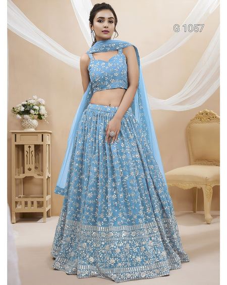 A woman models a light blue embroidered Indian ethnic wear set consisting of a sleeveless crop blouse, a long flared skirt, and a matching dupatta. The set features intricate white floral embroidery and is shown in a decorated indoor setting with beige walls, draped white fabric, and a wooden side table.