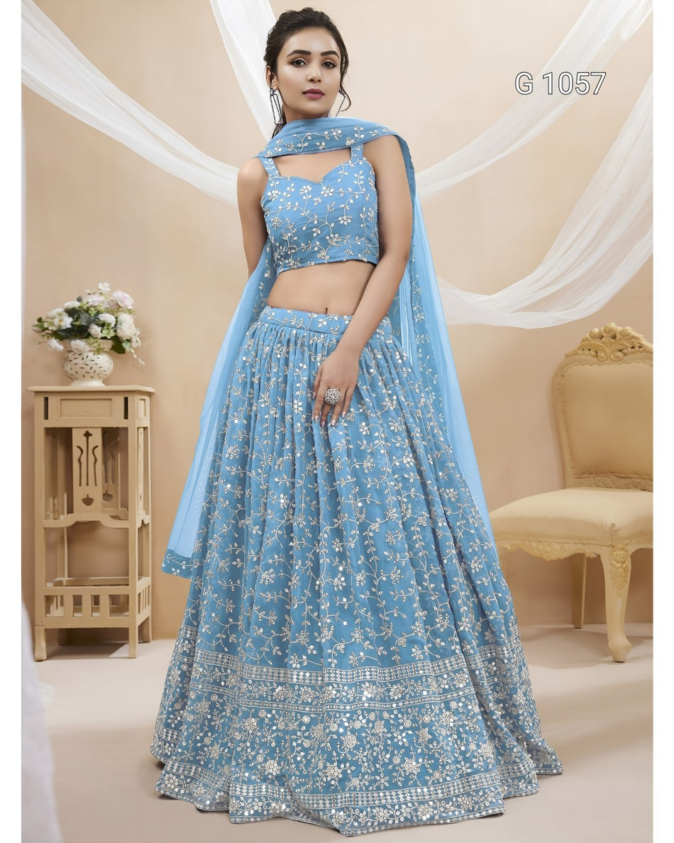 A woman models a light blue embroidered Indian ethnic wear set consisting of a sleeveless crop blouse, a long flared skirt, and a matching dupatta. The set features intricate white floral embroidery and is shown in a decorated indoor setting with beige walls, draped white fabric, and a wooden side table.