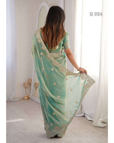 A woman viewed from behind wearing a mint green saree with gold floral embroidery and a matching embroidered border, standing in a bright room with white curtains. The text 'G 994' appears in the top right corner.