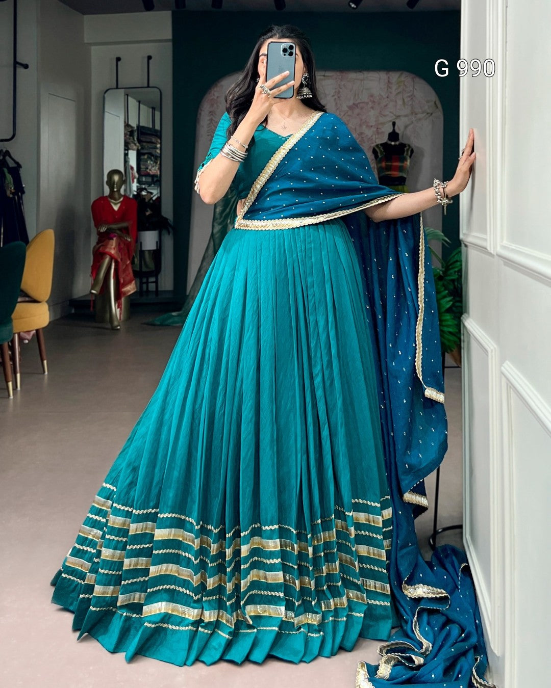 A woman takes a mirror selfie in a boutique wearing a teal blue traditional Indian dress with a matching dupatta. The dress features a pleated skirt with gold and beige horizontal stripes near the hem and a heavily embellished blue border.