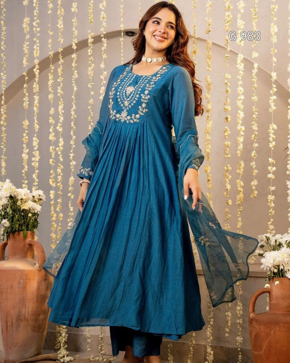 A woman models a teal blue long kurta with intricate silver embroidery on the chest, paired with matching blue trousers and a sheer blue dupatta, set against an arched backdrop decorated with hanging white flowers.