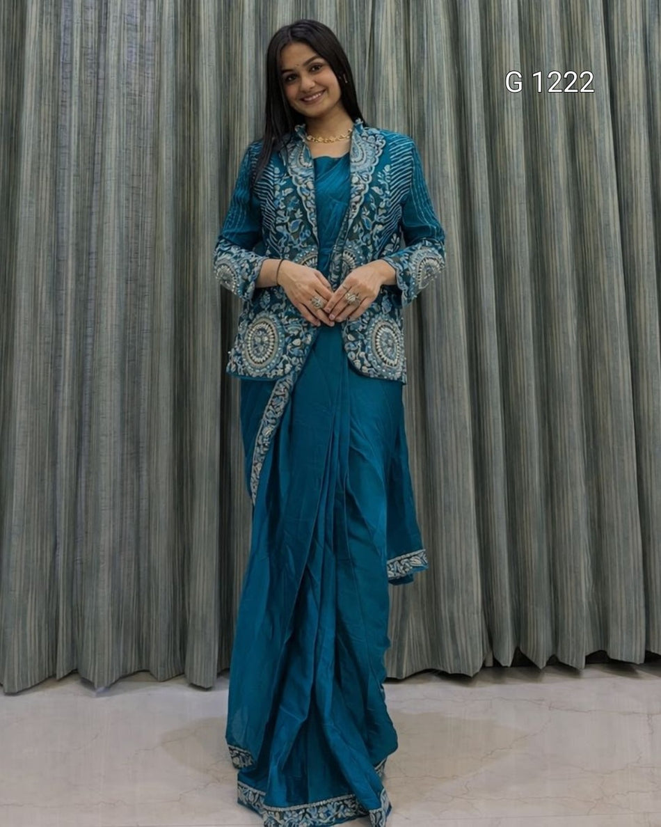 A woman modeling a teal blue traditional outfit with intricate silver embroidery, consisting of a kurta, matching skirt, and dupatta, set against a gray pleated curtain backdrop.