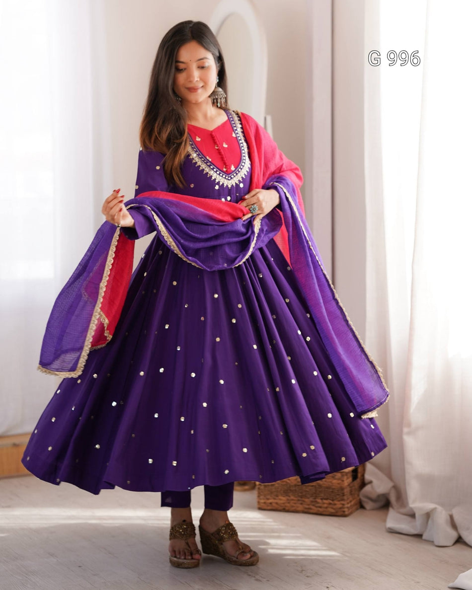 A woman modeling a traditional Indian outfit consisting of a purple flared skirt with gold star patterns, a red embroidered blouse, and a purple dupatta with red borders, set against a bright indoor background with white curtains.