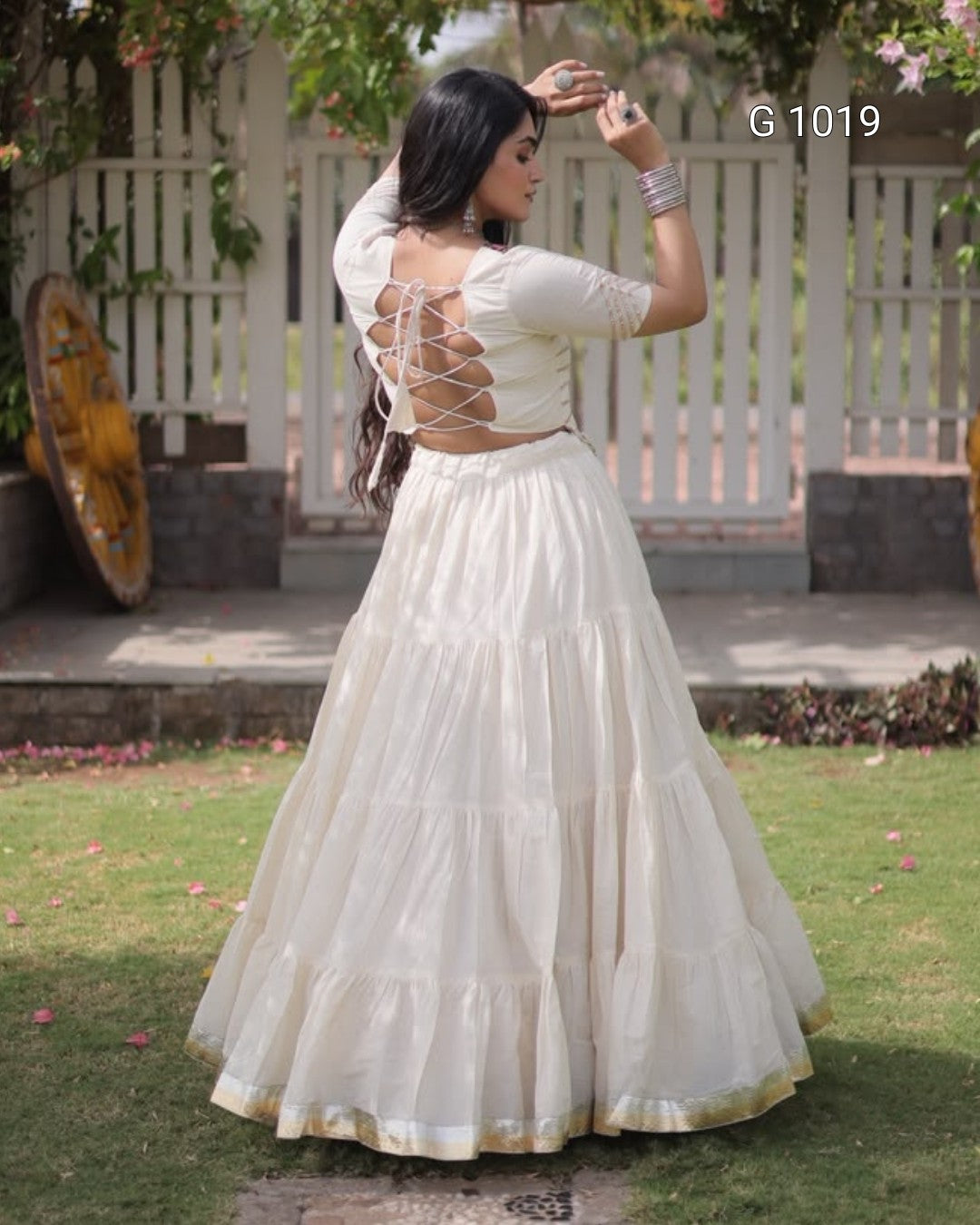 A woman wearing a cream-colored tiered Anarkali-style dress with lace-up back detailing, standing outdoors in a garden setting. The image includes the text overlay 'G 1019'.