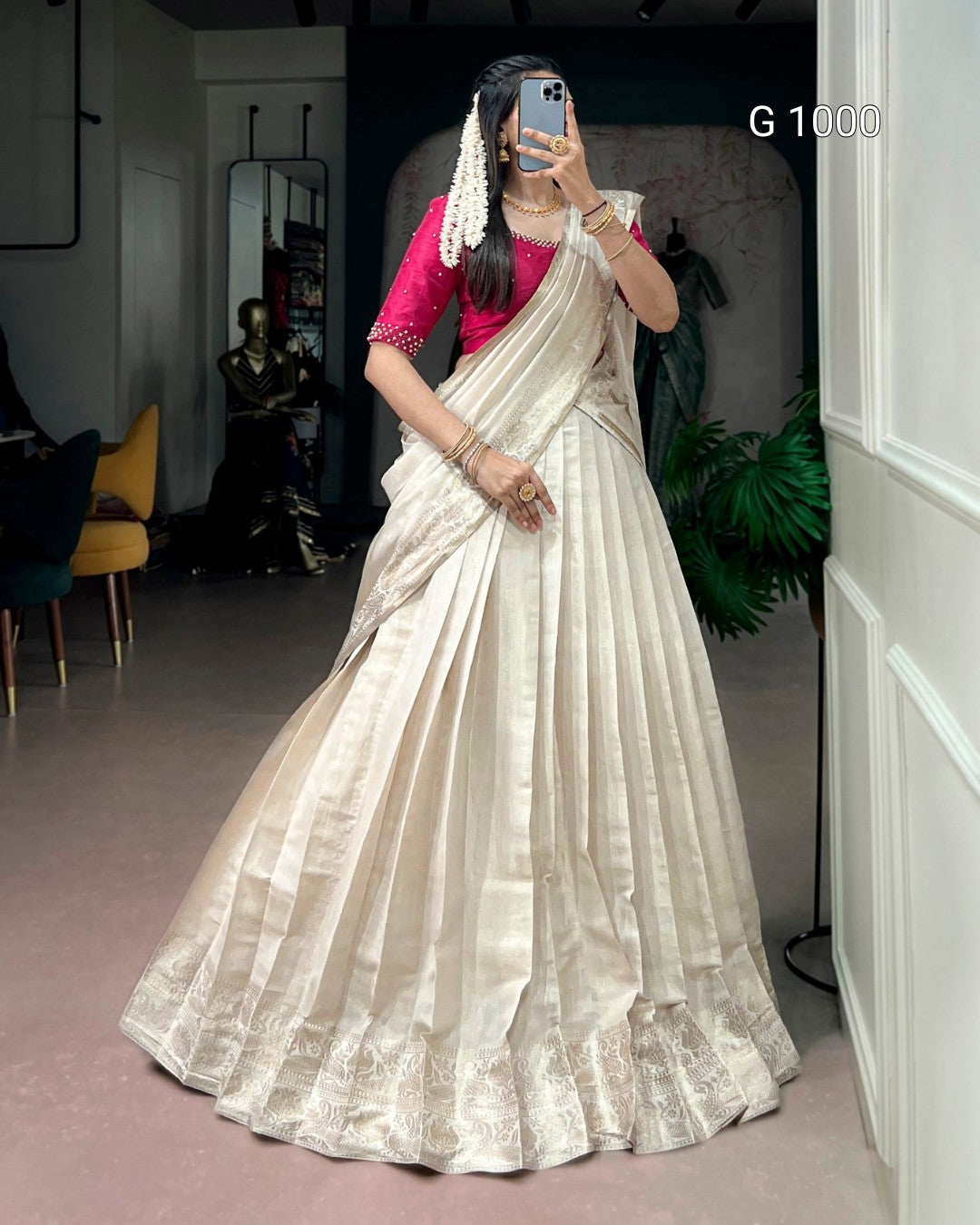 A woman takes a mirror selfie wearing a traditional Indian lehenga choli. The outfit consists of a voluminous cream-colored silk saree with a heavily embroidered border and a bright pink sleeveless blouse.