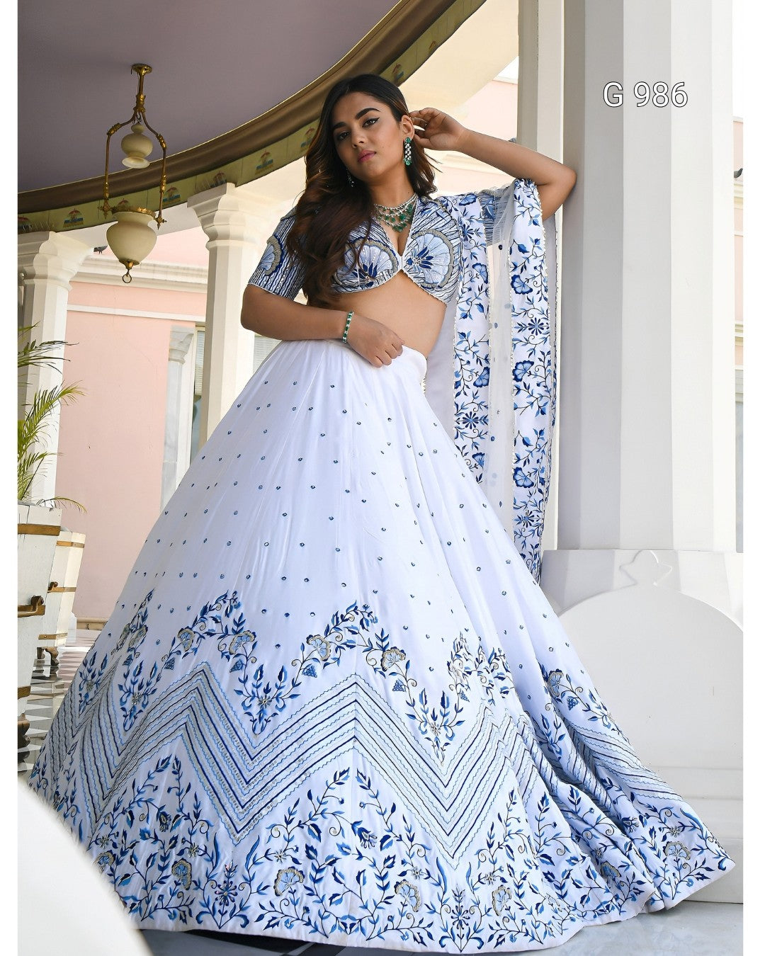 A woman modeling a white and blue printed lehenga choli set in an elegant indoor architectural setting with white pillars. The outfit features a heavily embellished blue floral and geometric border on the flared skirt, a matching printed blouse, and a coordinating dupatta.
