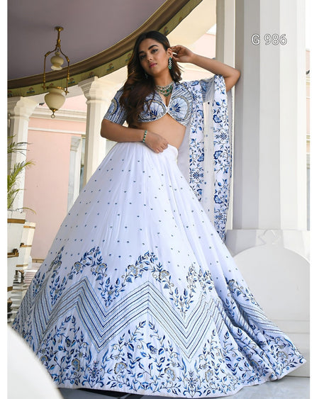 A woman modeling a white and blue printed lehenga choli set in an elegant indoor architectural setting with white pillars. The outfit features a heavily embellished blue floral and geometric border on the flared skirt, a matching printed blouse, and a coordinating dupatta.
