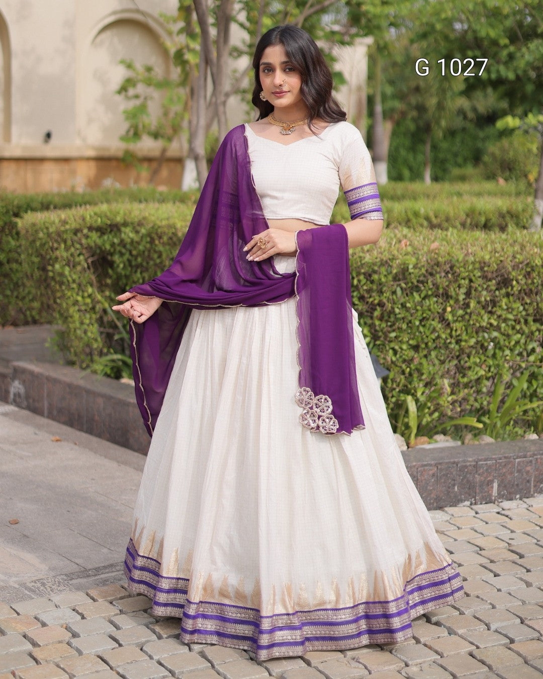 A woman modeling a traditional Indian ethnic wear set featuring an off-white/cream Anarkali skirt with purple and gold borders, a matching cream blouse with purple sleeve bands, and a deep purple dupatta draped over her shoulder.
