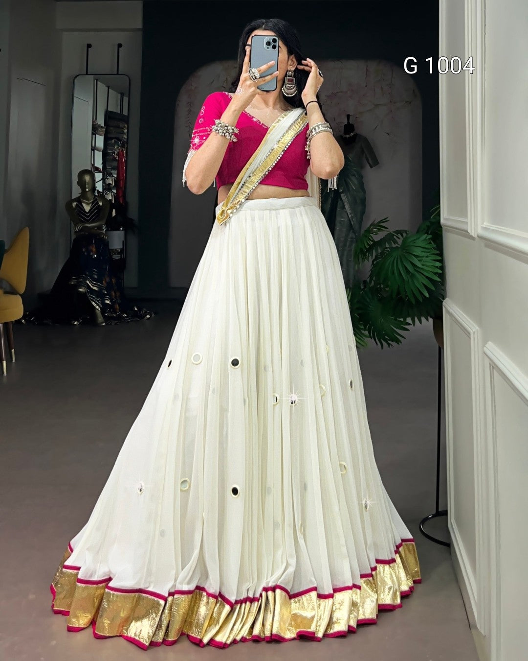 A woman takes a mirror selfie wearing a traditional Indian outfit consisting of a white flared skirt with gold and pink borders, a bright pink crop top, and a gold and pink draped scarf. The background is a dark room with a mirror, plant, and furniture.