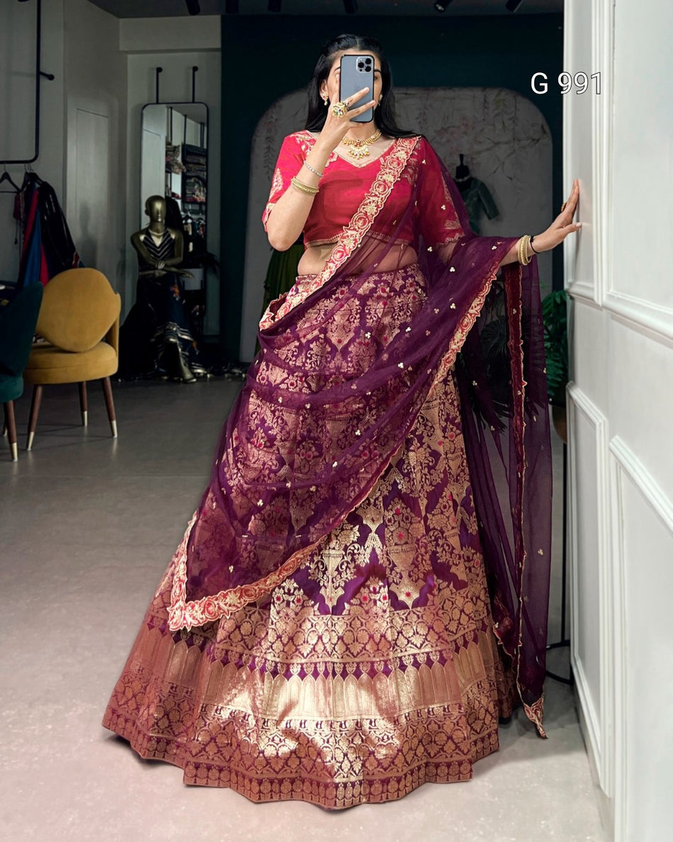 A woman takes a mirror selfie wearing an ornate Indian lehenga choli set. The outfit features a deep purple dupatta and skirt with intricate gold zari embroidery, paired with a bright pinkish-red blouse.