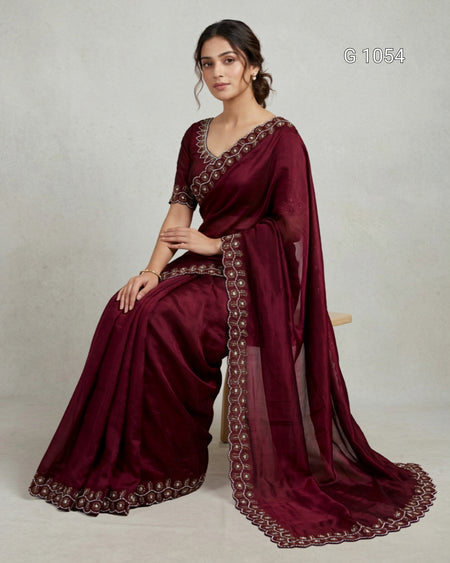 A woman seated on a small stool wearing a deep maroon saree with ornate gold embroidered borders and a matching sheer dupatta, photographed against a plain light gray studio background.