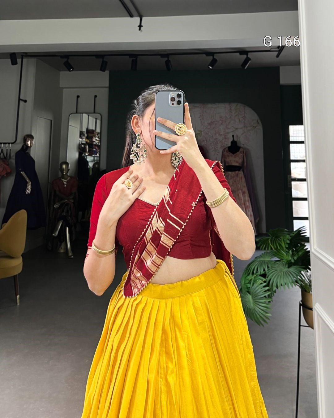 Woman taking a mirror selfie wearing a red blouse and yellow saree in a store setting.