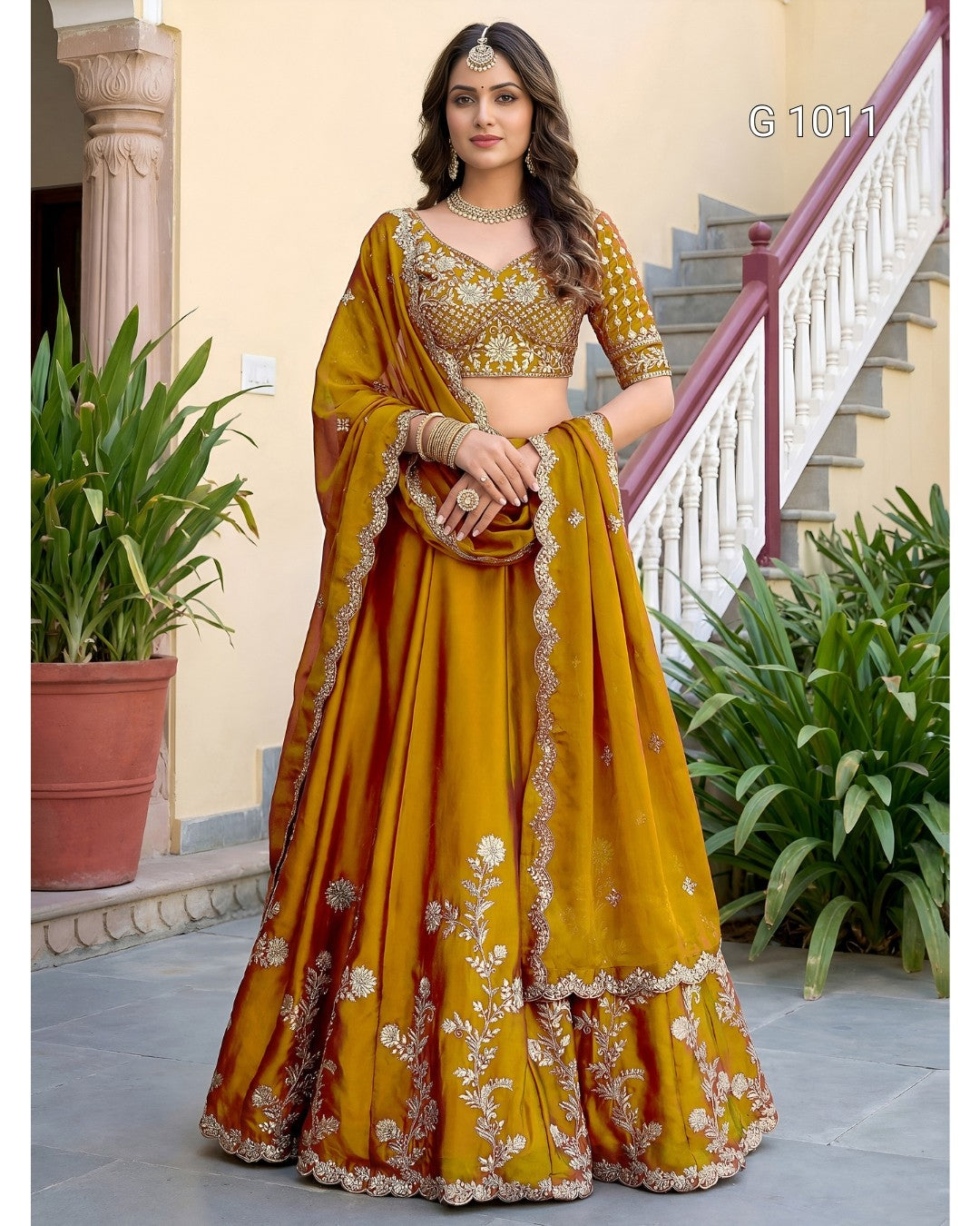 A woman models a traditional Indian lehenga choli set in mustard yellow with intricate gold and silver floral embroidery. The set includes a heavily embroidered choli, a full flared skirt (lehenga), and a dupatta draped over her arm.