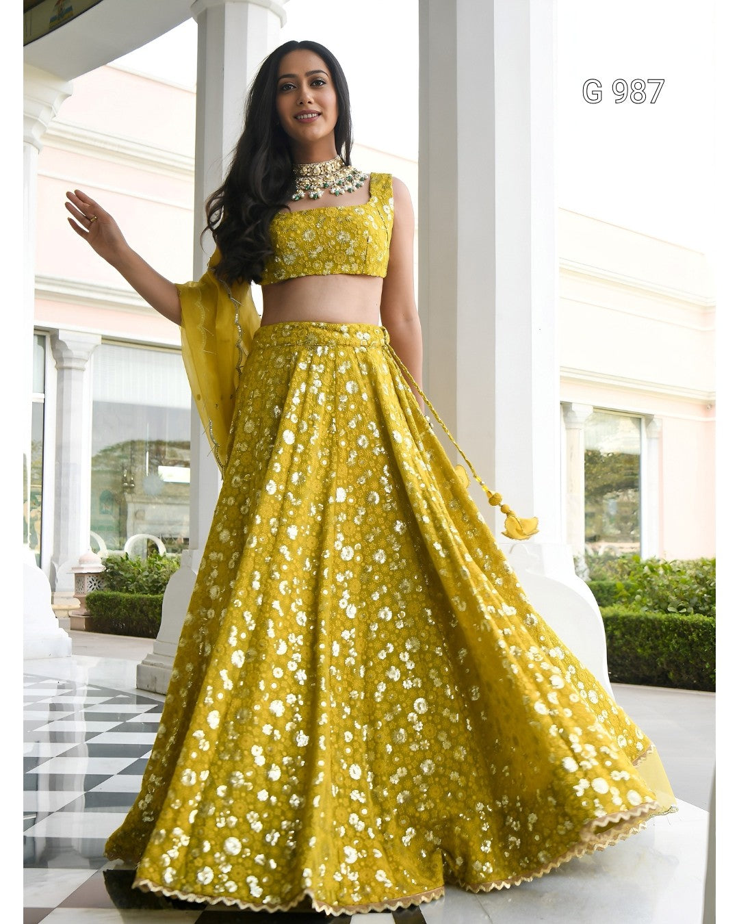 A woman modeling a bright yellow embroidered lehenga choli set with a matching dupatta in an outdoor colonnade. The set features intricate white floral embroidery and mirror work.