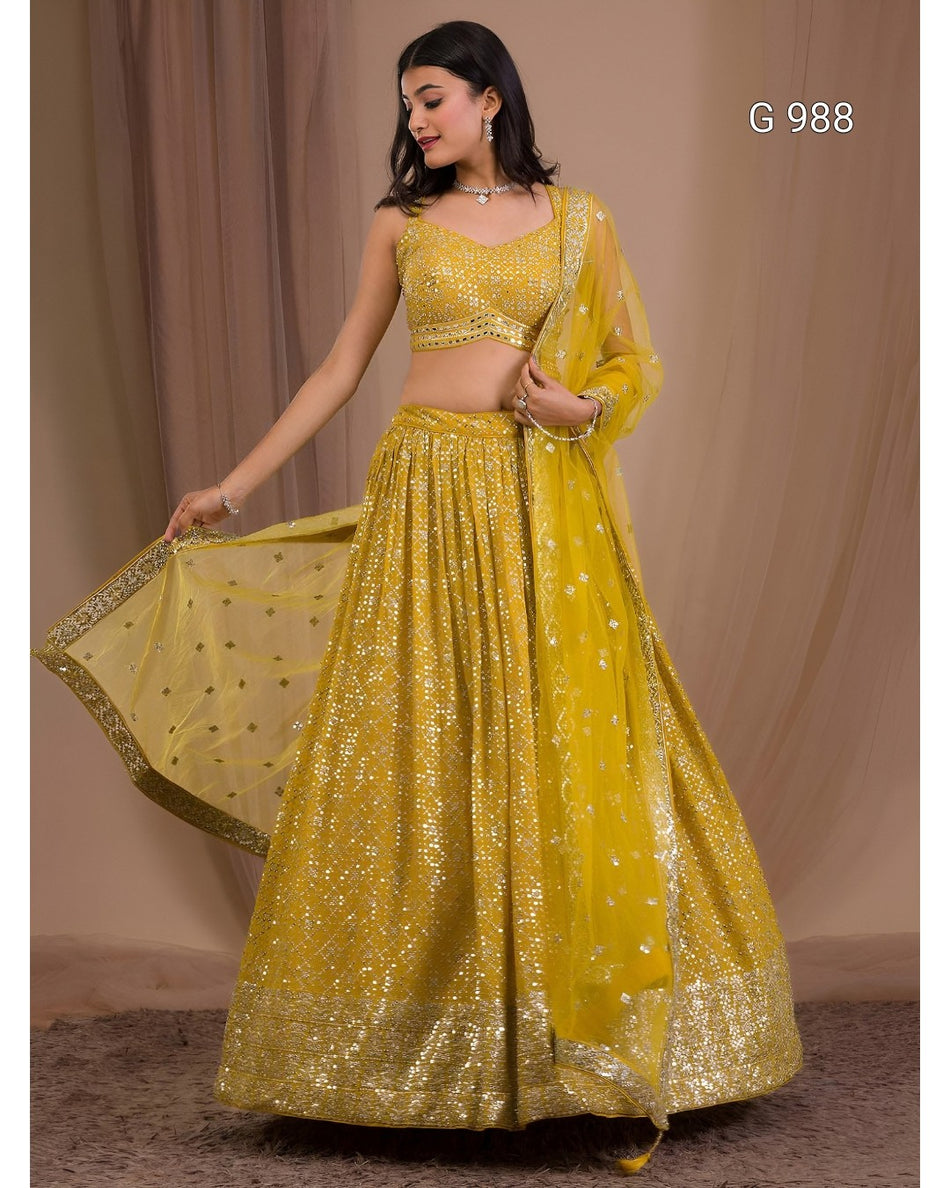A woman models a bright yellow embellished lehenga choli set with a matching dupatta, standing against a beige curtain backdrop. The set features intricate gold sequin and mirror work, and the text 'G 988' is visible in the top right corner.