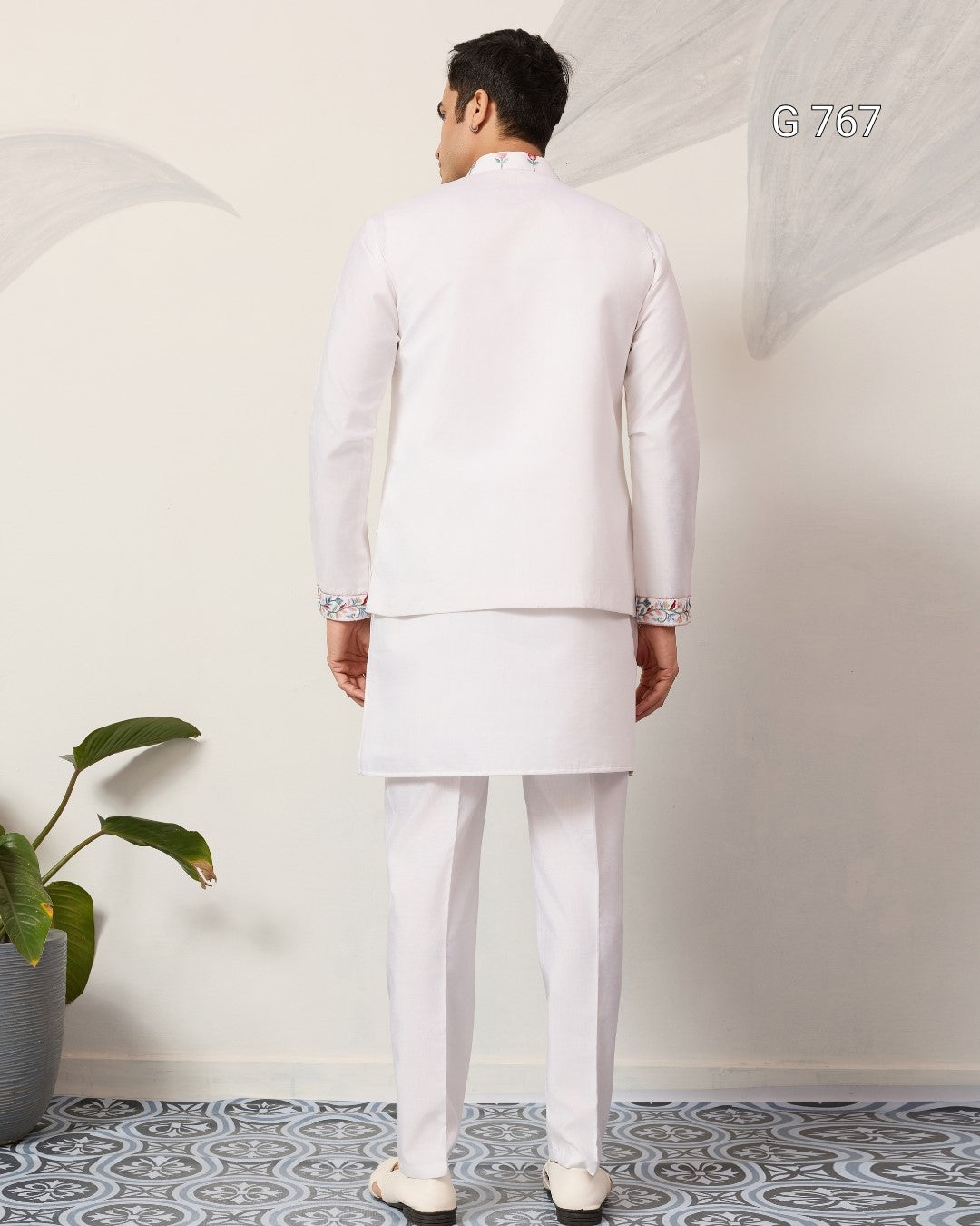 Man wearing a white traditional outfit standing against a white wall with a plant on the left.