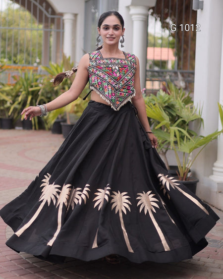 A woman modeling a black traditional skirt with embroidered beige palm tree patterns and a heavily embellished multicolor embroidered bodice, set against an outdoor courtyard background.