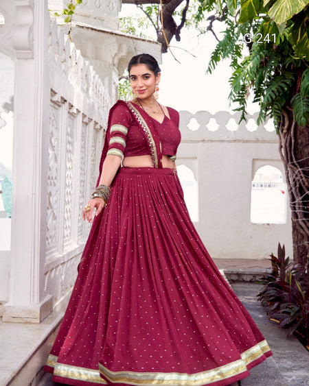 Woman in a maroon traditional outfit standing in an outdoor setting with white architecture and greenery.