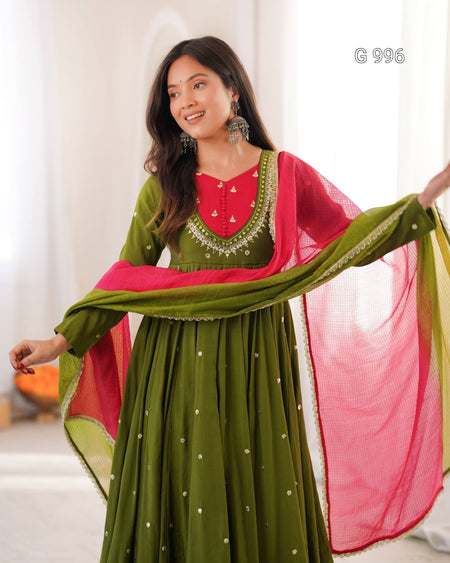 A woman modeling a traditional Indian outfit featuring an olive green anarkali skirt with small gold embroidered dots, a bright red V-neck inner blouse with gold embroidery, and a matching pink dupatta draped over her arm.