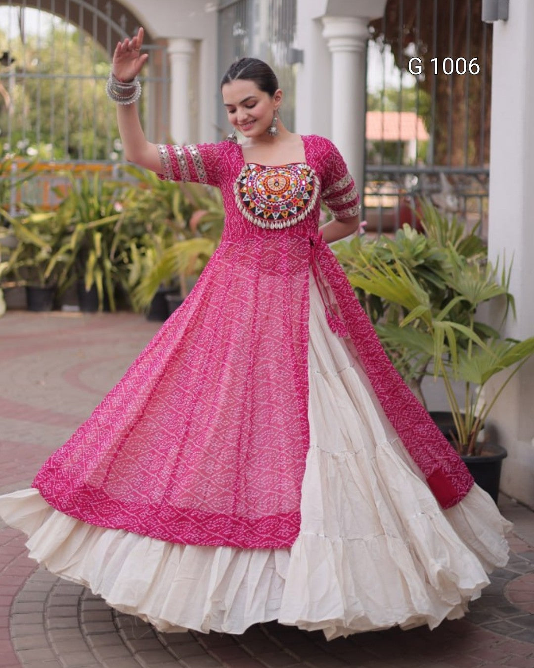 A woman modeling a traditional Indian anarkali gown with a bright pink embroidered bodice and sleeves, a matching pink dupatta, and a tiered off-white skirt, posed in an outdoor courtyard setting.