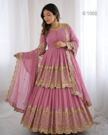 A woman modeling a pink traditional Anarkali gown with gold embroidery and a matching sheer dupatta, set against a white curtain background.