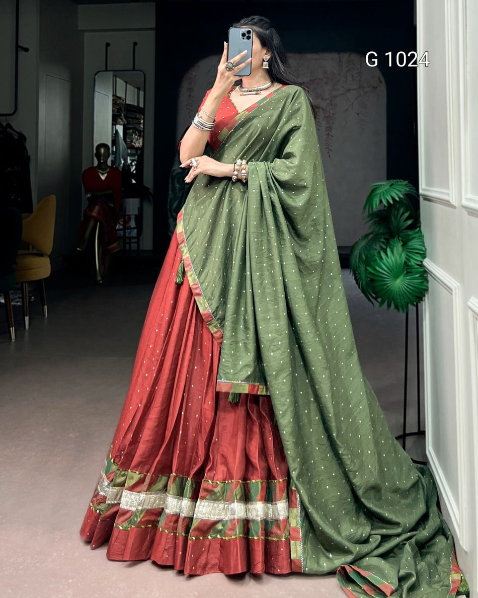 A woman takes a mirror selfie wearing a traditional Indian outfit consisting of a rust-red flared skirt (lehenga) with a decorative gold and green border, paired with a sage green saree drape (pallu) featuring small gold motifs. The text 'G 1024' is visible in the top right corner.