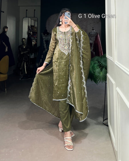 Woman wearing an olive green traditional outfit with embroidery, standing in a room.