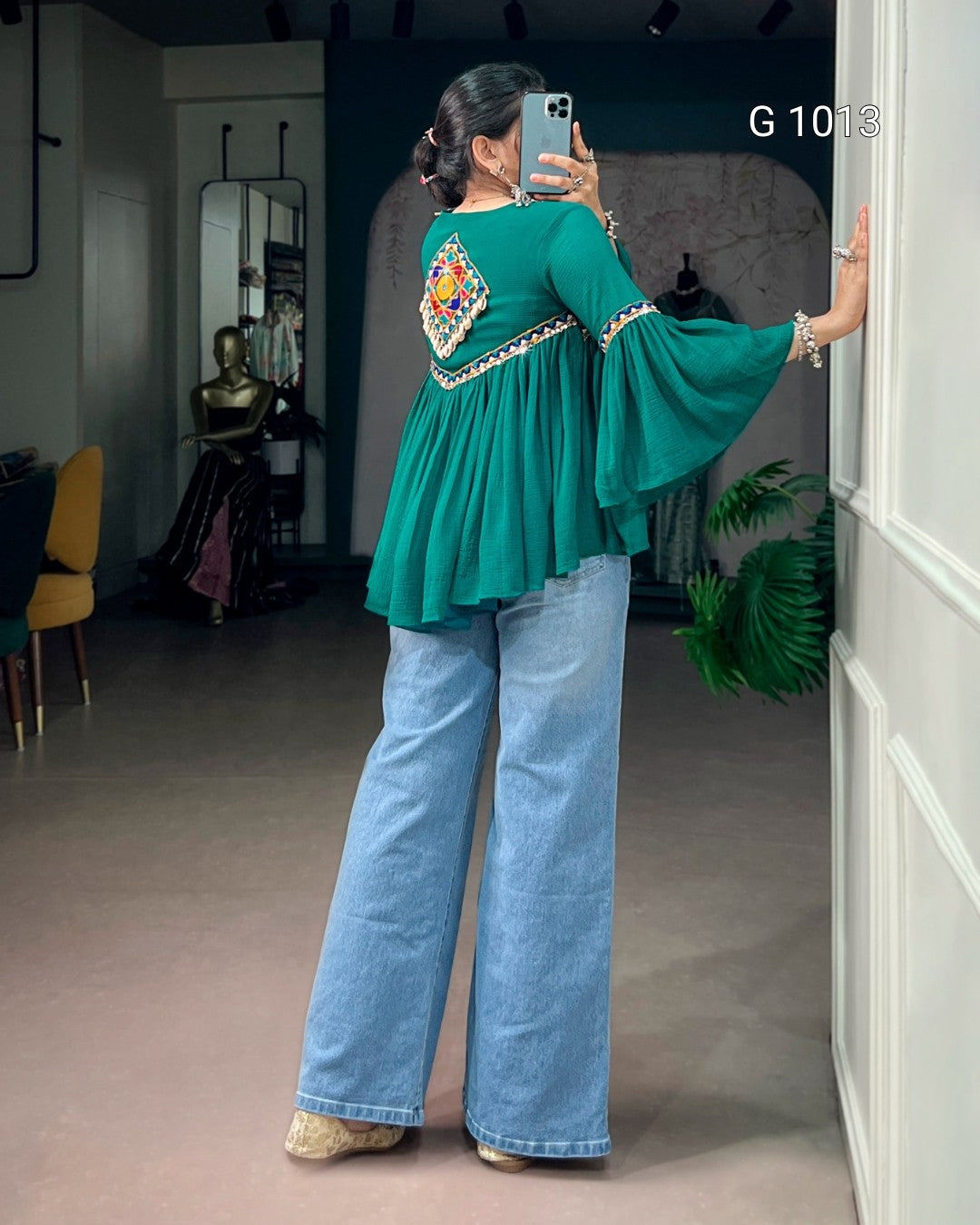A woman takes a mirror selfie in a boutique, wearing a teal green long-sleeve blouse with a colorful embroidered yoke and wide bell sleeves, paired with light-wash wide-leg jeans.
