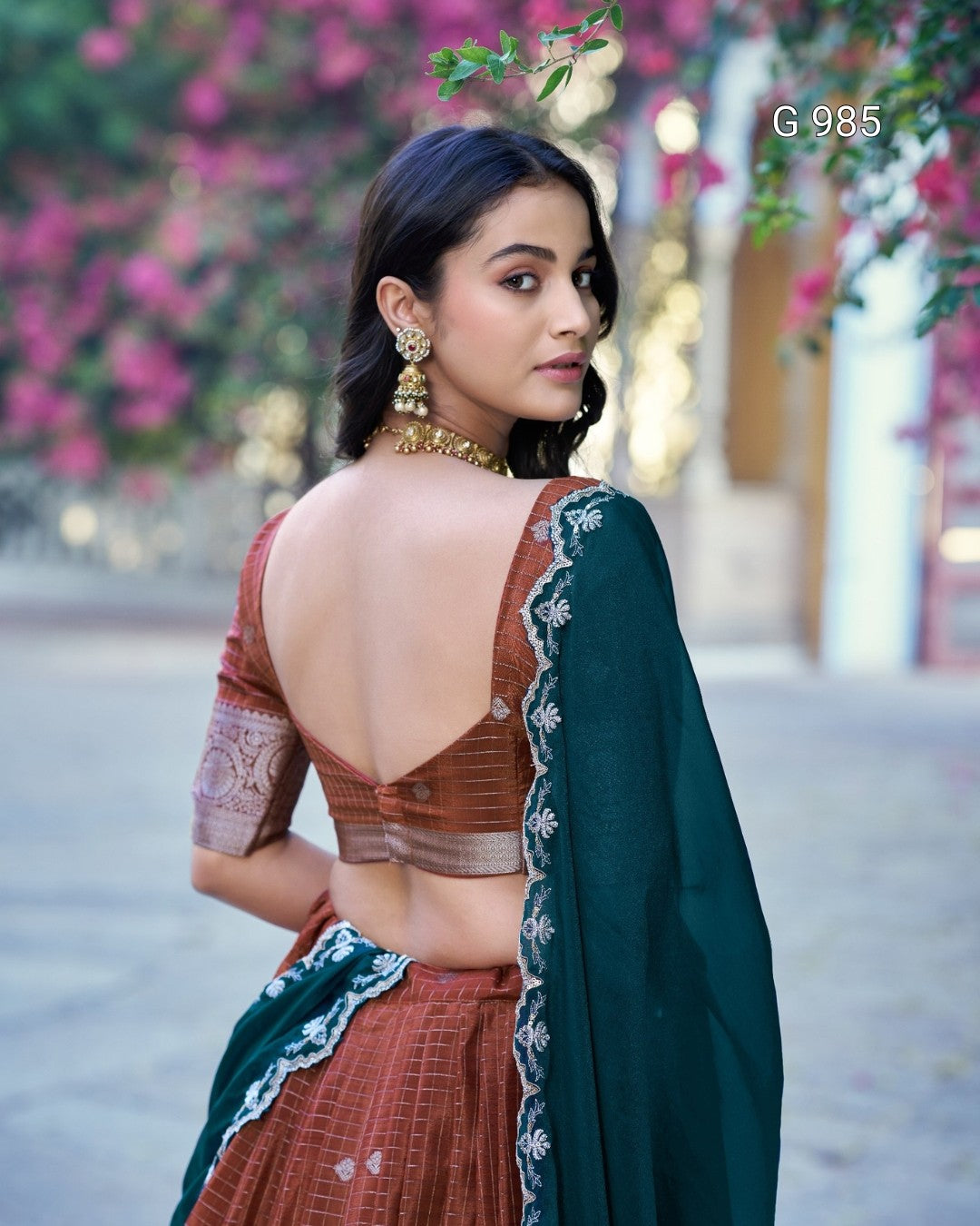 A woman wearing a traditional Indian outfit with a rust-colored embroidered blouse, matching skirt, and a dark teal dupatta, photographed outdoors with pink flowers in the background.