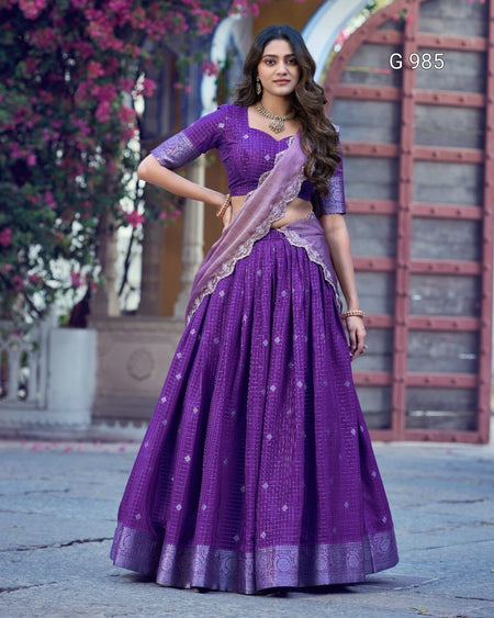 A woman modeling a purple traditional Indian outfit with a matching skirt, blouse, and dupatta, standing outdoors in front of a stone and wood structure with pink flowers on the left. The number 'G 985' appears in the top right corner.