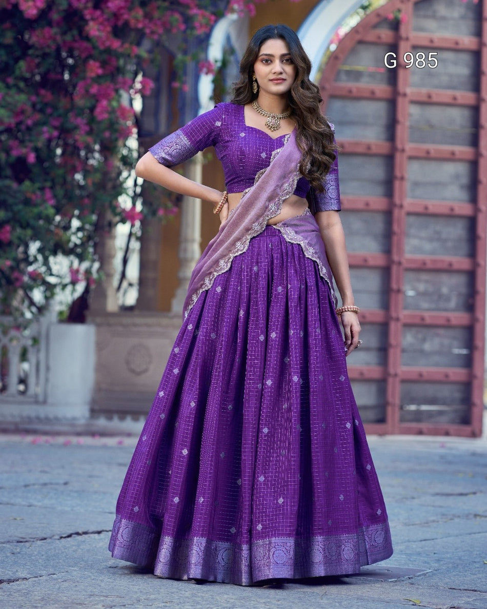 A woman modeling a purple traditional Indian outfit with a matching skirt, blouse, and dupatta, standing outdoors in front of a stone and wood structure with pink flowers on the left. The number 'G 985' appears in the top right corner.