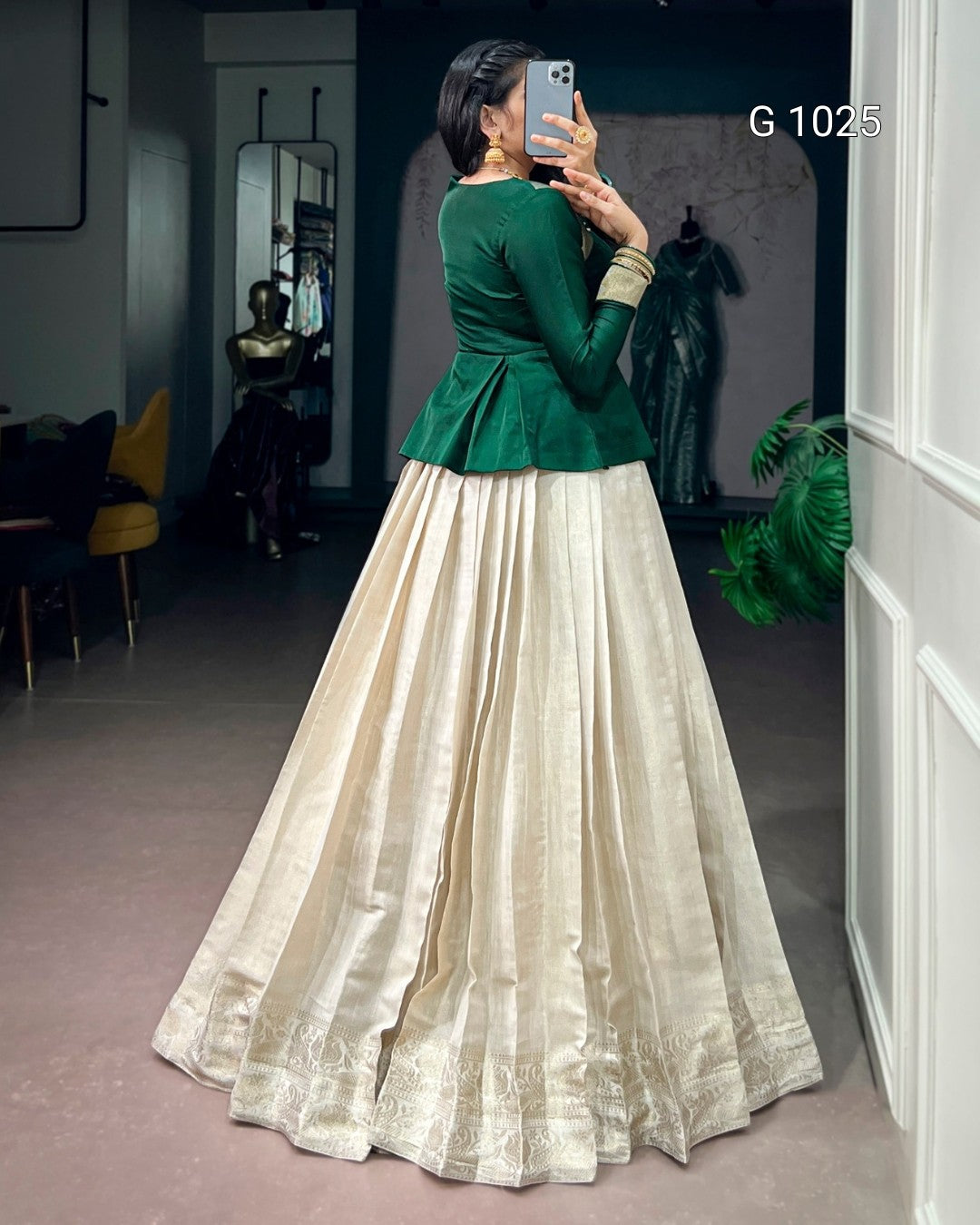 A woman takes a mirror selfie in a boutique wearing a traditional Indian outfit consisting of a dark green velvet peplum top and a voluminous cream-colored silk Anarkali skirt with gold zari embroidery.