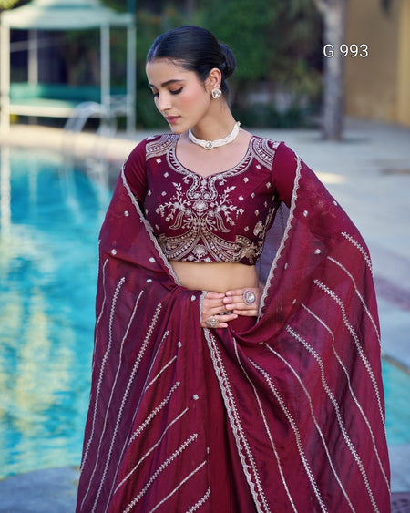 A woman models a deep maroon traditional Indian outfit with intricate gold embroidery, standing next to a swimming pool. The outfit consists of a heavily embellished crop top and a matching draped skirt or dupatta with gold vertical stripes.