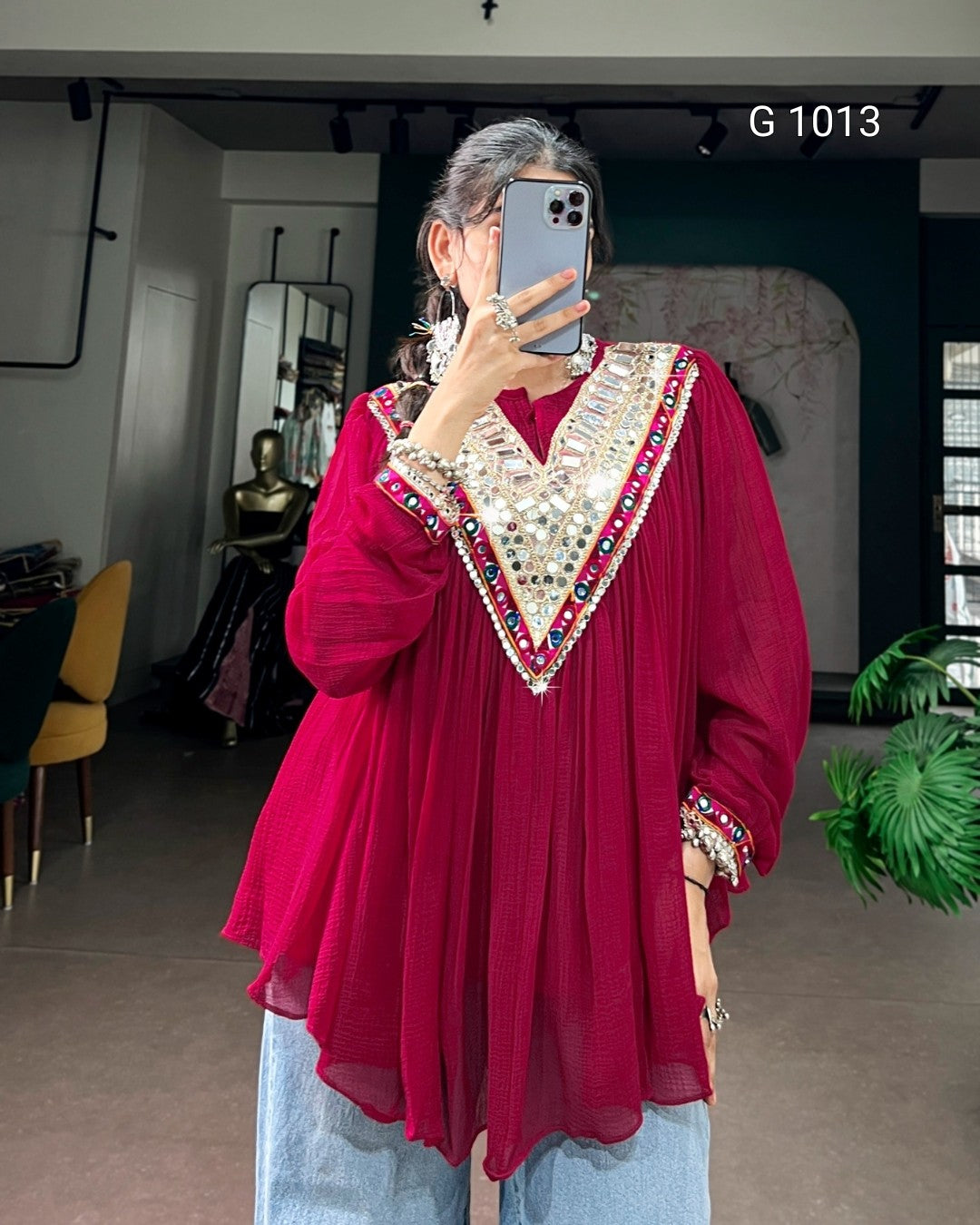 A woman takes a mirror selfie wearing a long-sleeved magenta-red tunic top with a heavily embellished V-neckline and cuffs in silver, red, and blue sequins, paired with light blue jeans.