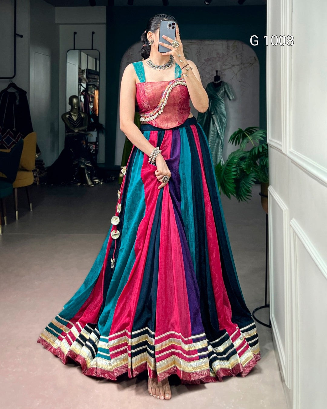 A woman takes a mirror selfie in a boutique wearing a traditional Indian lehenga choli. The dress features a metallic pink choli and a flared skirt with vertical stripes of teal, magenta, purple, and black, finished with a decorative gold and white border.