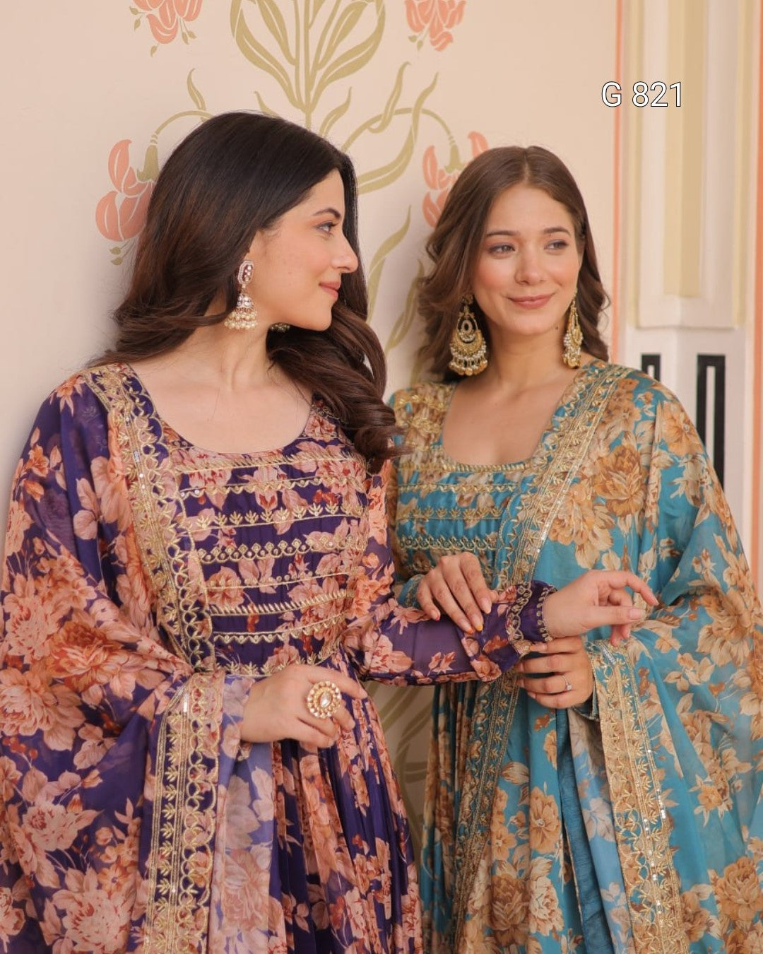 Two women in traditional outfits with floral patterns standing against a decorative wall.