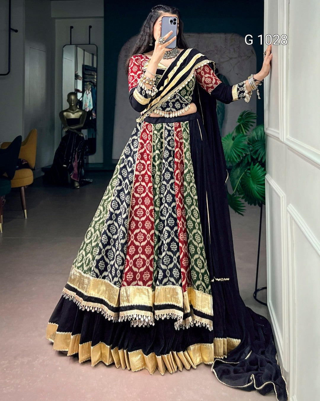 A woman takes a mirror selfie in a boutique wearing an ornate traditional Indian lehenga choli with a black dupatta. The outfit features intricate red, gold, and black patterns on the skirt, with a red and black embroidered bodice and sleeves.