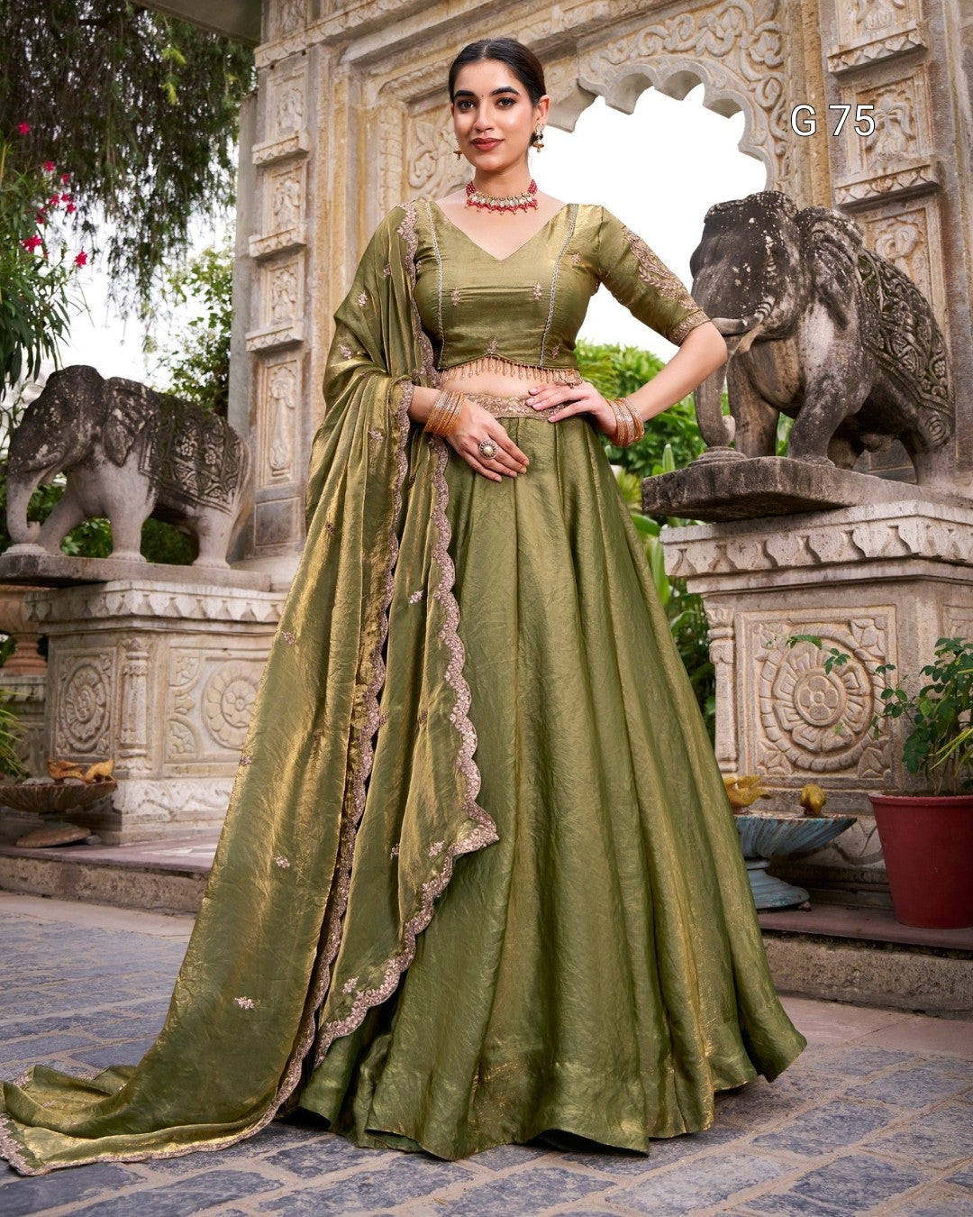 A woman models an olive green traditional Indian lehenga choli suit with a matching dupatta, standing in front of an ornate stone archway decorated with elephant statues.
