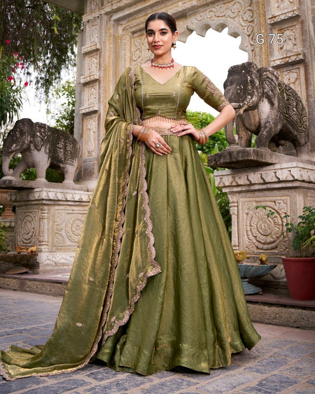 A woman models an olive green traditional Indian lehenga choli suit with a matching dupatta, standing in front of an ornate stone archway decorated with elephant statues.