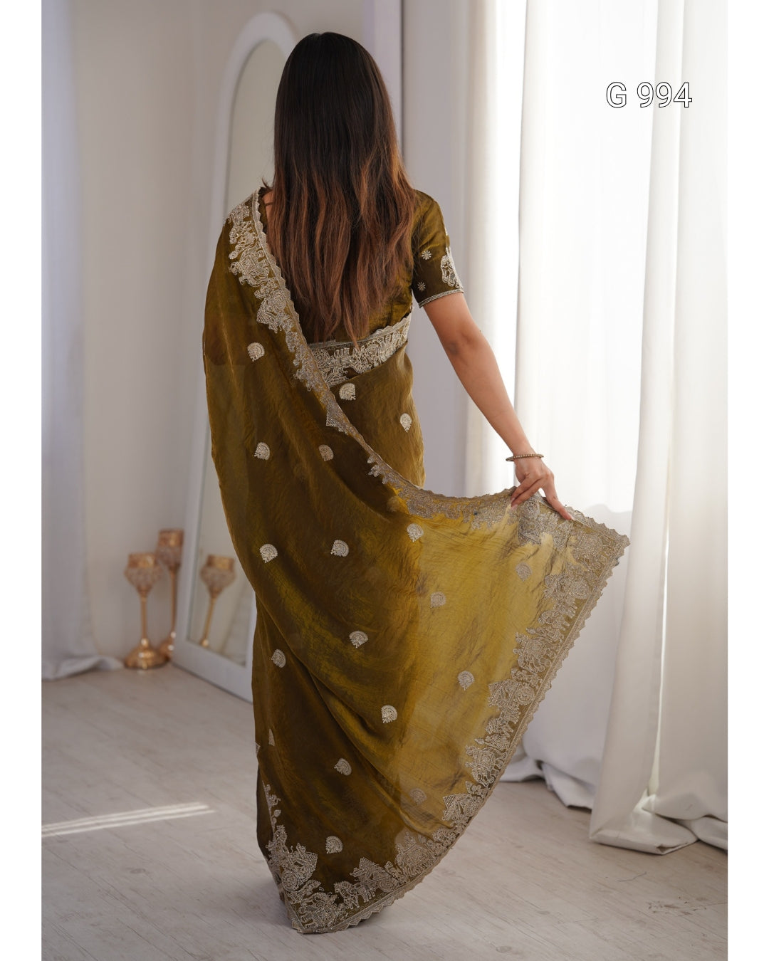 A woman viewed from behind wearing an olive green saree with beige floral embroidery and a matching embroidered border, holding out the fabric in a bright indoor setting.