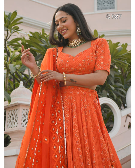 A woman wearing a bright orange embroidered traditional dress with a matching sheer dupatta, standing outdoors in front of a light pink building and green plants.