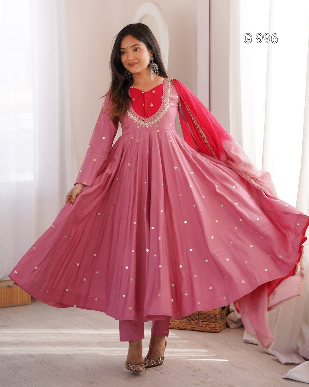 A woman modeling a pink traditional Anarkali gown with a red embroidered neckline, silver mirror work, matching pink trousers, and a red dupatta with silver mirror work, set against a soft indoor background with white curtains.