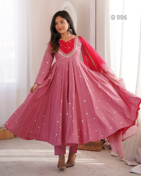 A woman modeling a pink traditional Anarkali gown with a red embroidered neckline, silver mirror work, matching pink trousers, and a red dupatta with silver mirror work, set against a soft indoor background with white curtains.
