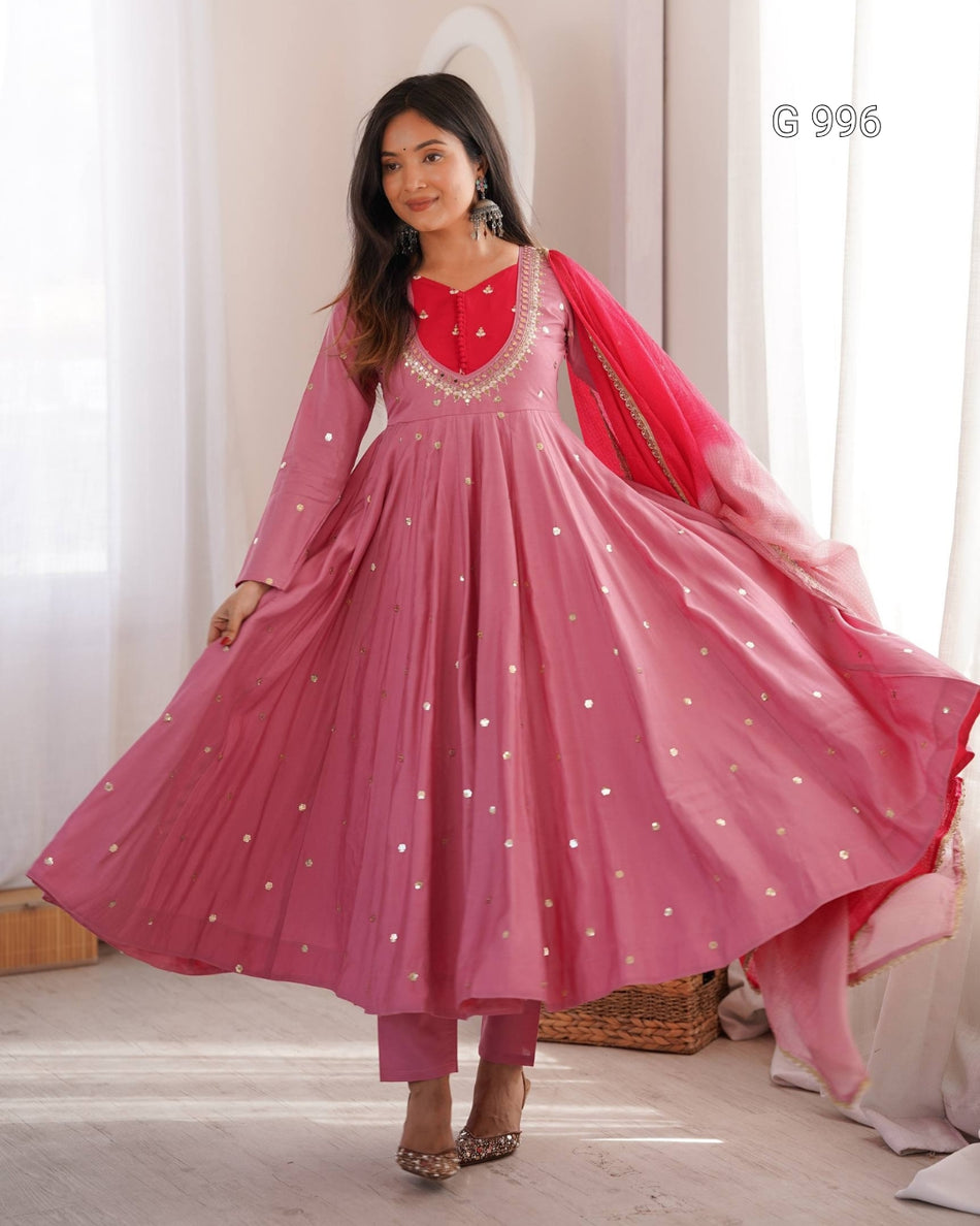 A woman modeling a pink traditional Anarkali gown with a red embroidered neckline, silver mirror work, matching pink trousers, and a red dupatta with silver mirror work, set against a soft indoor background with white curtains.