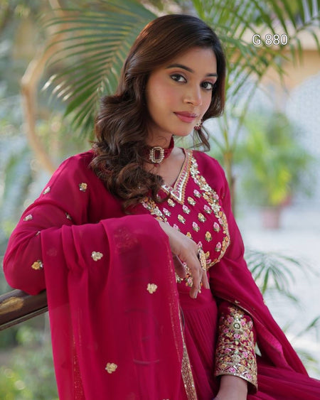 Woman in a red traditional outfit with gold embroidery, standing outdoors with greenery in the background.