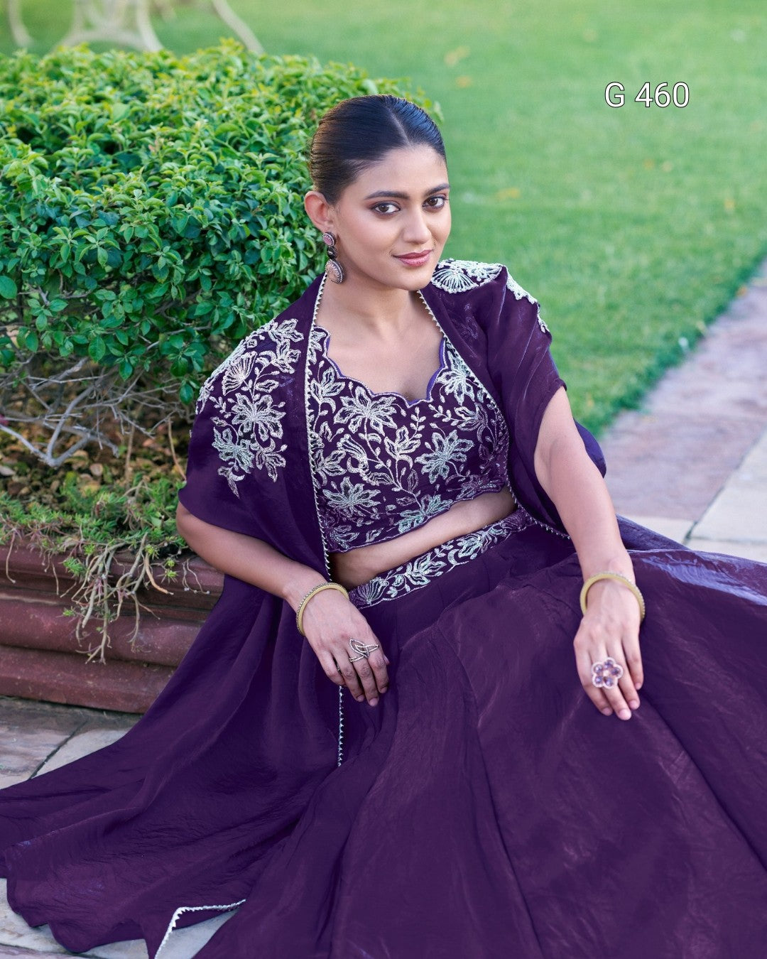 A woman seated outdoors on a stone path wearing a deep purple traditional outfit with silver floral embroidery on the blouse and draped shawl. Green shrubs and grass are in the background, and the text 'G 460' appears in the upper right.