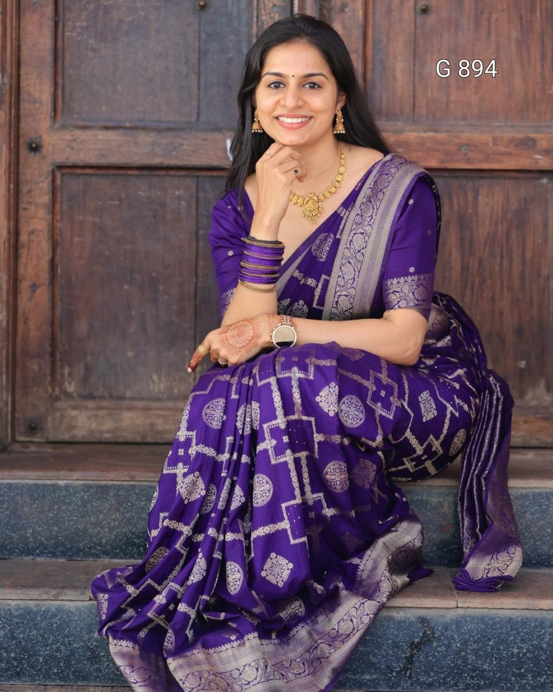 Woman in a purple saree sitting on steps with a wooden door in the background