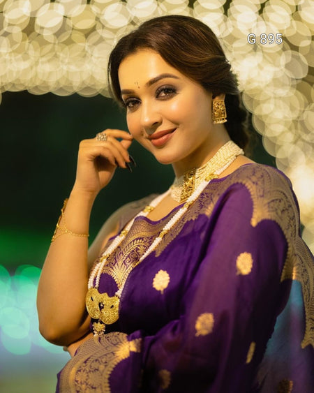 Woman in a purple saree with gold patterns against a blurred decorative background