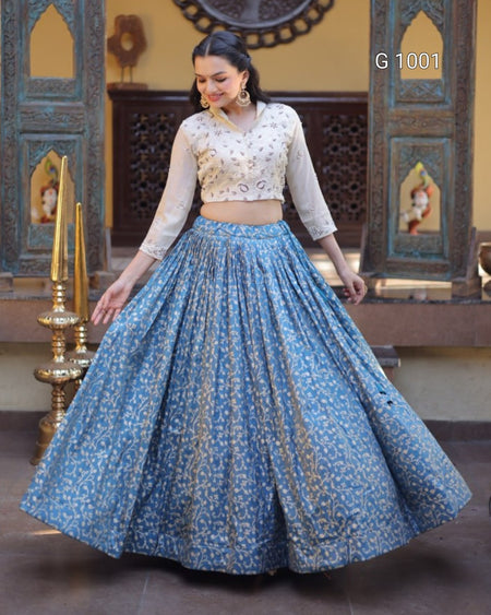 A woman modeling a traditional Indian ethnic wear set featuring a light blue floral printed skirt and a cream-colored embroidered crop top, set against an ornate architectural background.